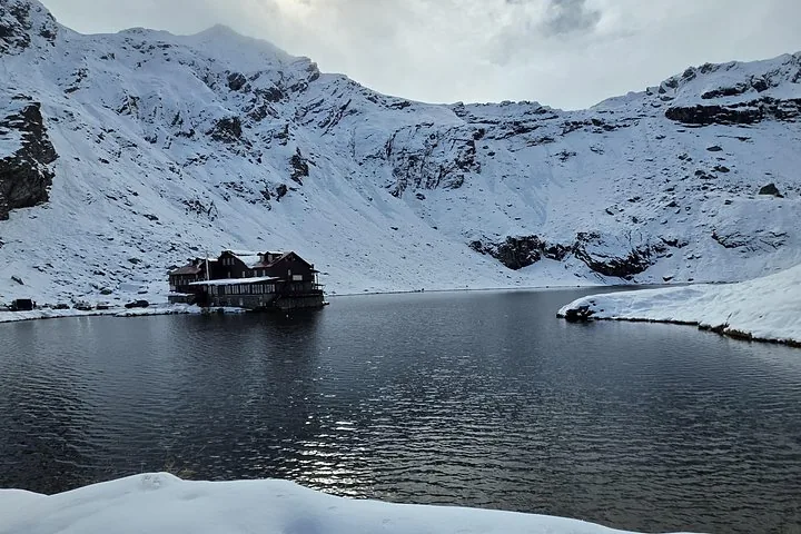 Eine Hütte an einem dunklen See, umgeben von schneebedeckten Bergen.